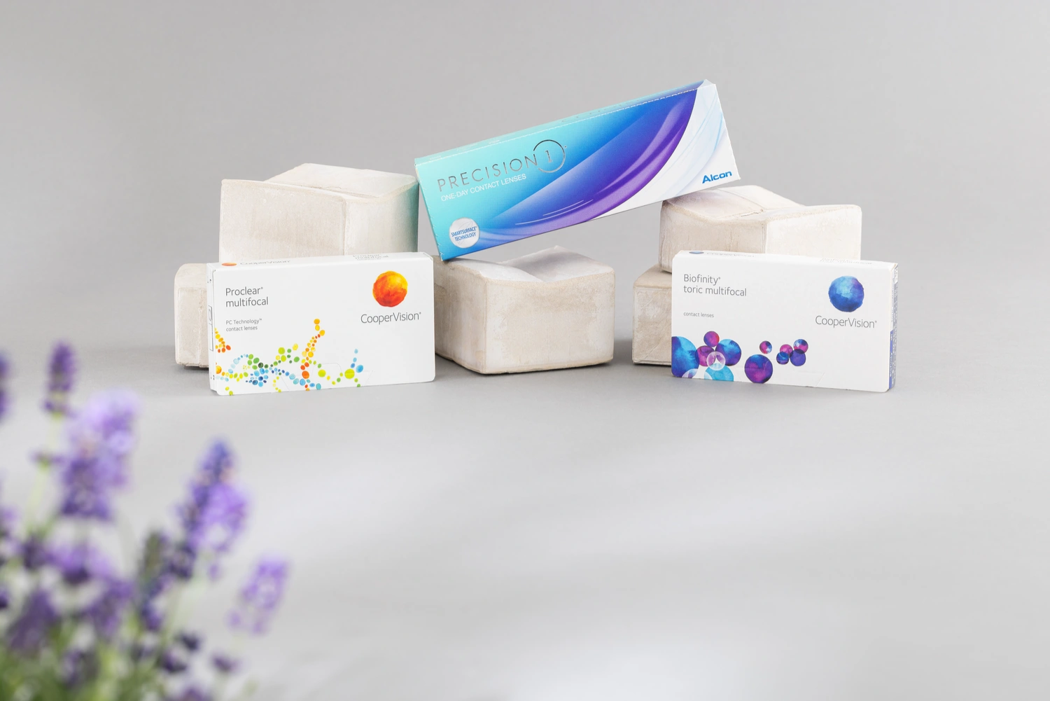 Drei verschiedene Kontaktlinsen-Boxen arrangiert mit einer lila Blume im Vordergrund.